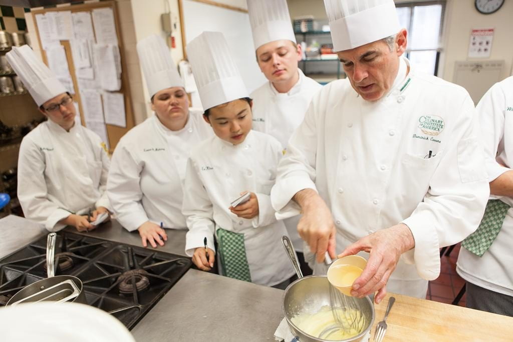 Chef Cerrone whisks egg into a mixture while five students in chef jackets observe in teh Culinary Immersion class.